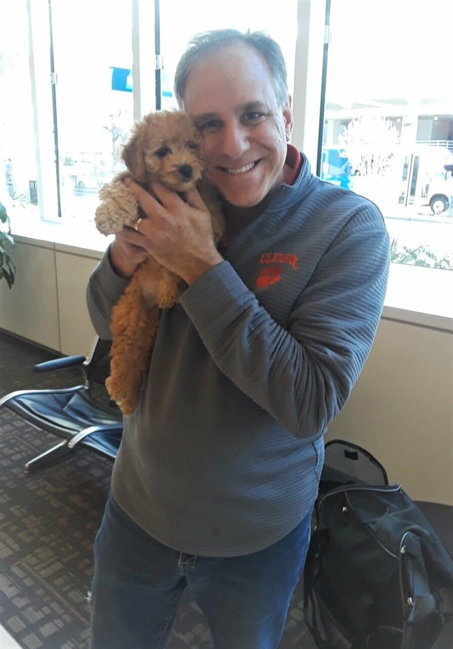 Barry Formanack, the president of Crockett Doodles, at the airport ready to fly with Lila the mini Goldendoodle in his lap to meet Bret Baier and his family and give them their new puppy.